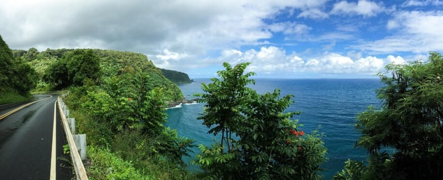 The Road to Hāna curves beside lush green cliffs overlooking the blue Pacific Ocean on Maui.