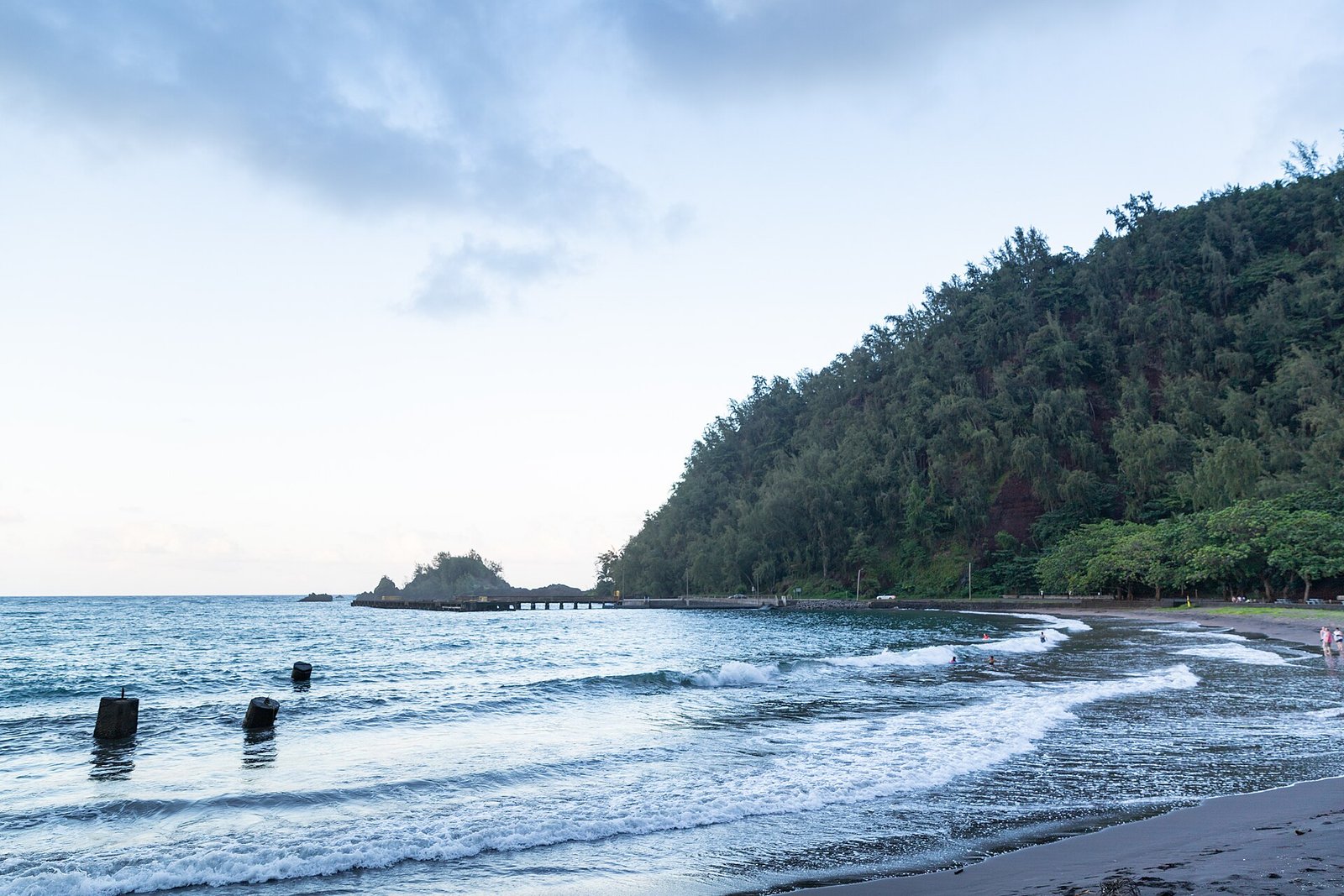 Hāna Bay beach with the pier and green hillside in the distance